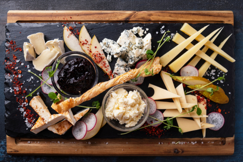 Na imagem, há uma tábua de frios com queijo parmesão e outros tipos de queijo, grissini e geleias