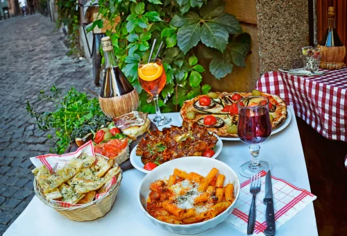 Na imagem, há uma mesa ao ar livre com vinho e pratos com comidas italianas