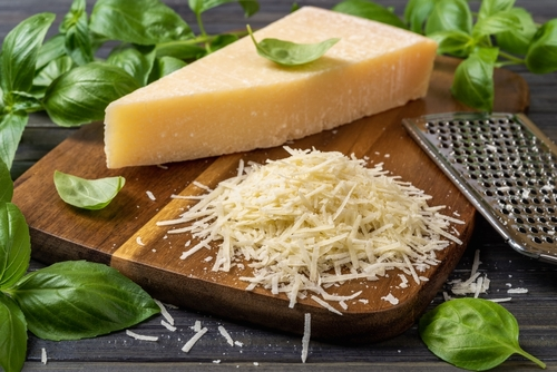 Na imagem, há um queijo parmesão em peça ao lado de queijo parmesão ralado em tábua de madeira