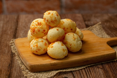 Na imagem, há uma pilha de pão de queijo em cima de tábua de madeira