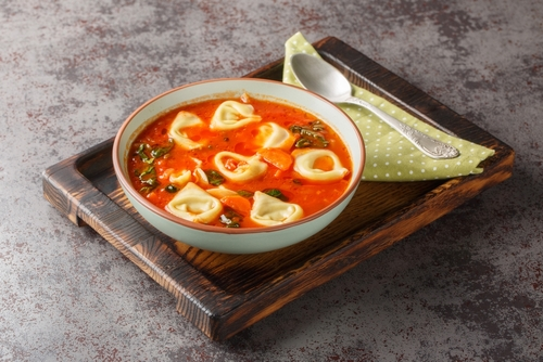 Na imagem, há uma tigela com sopa de tomate e tortellini em cima de bandeja