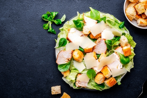 Na imagem, há um prato de salada ceaser com alface, pedaços de frango e croutons